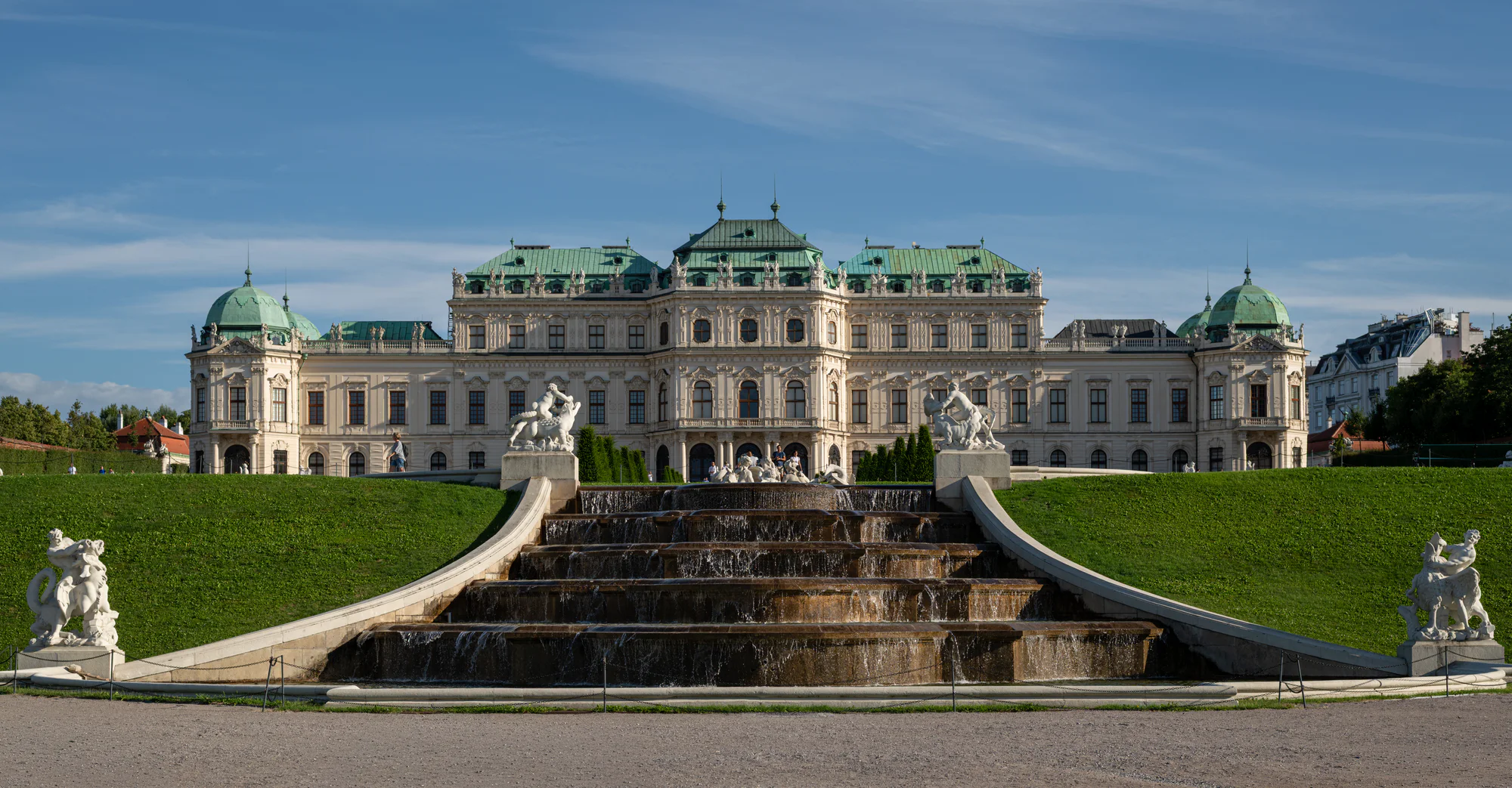 Upper Belvedere’s pale façade and green roofs above a stepped garden cascade, with stone sculptures on both sides and a blue sky.