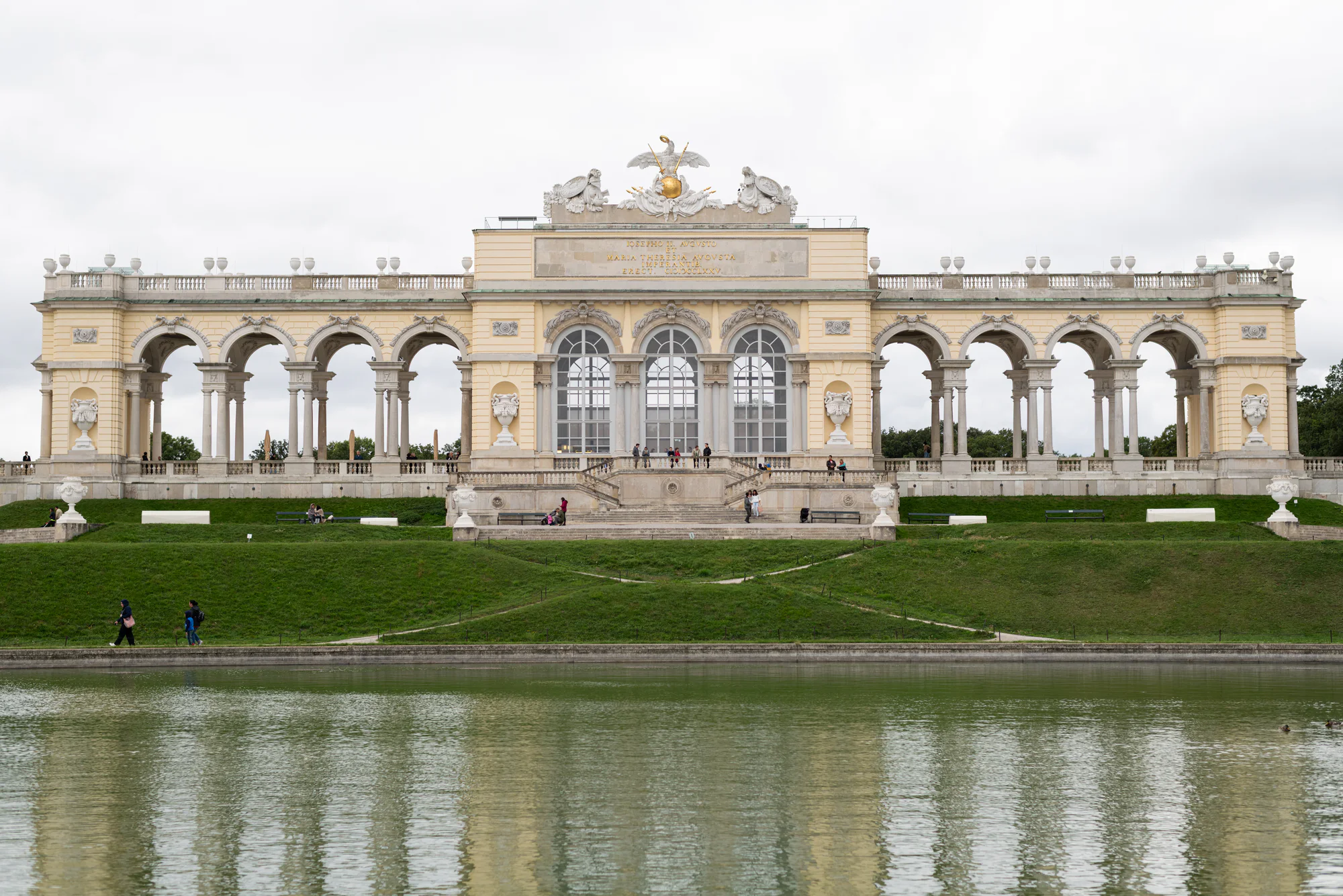The Gloriette’s pale yellow arcades and central hall above a green slope and reflecting pool, with visitors on the paths and terrace.
